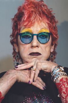 Alt text: "Portrait of Judy Chicago, an iconic Jewish American artist, known for her bold, feminist artwork. She is depicted with striking red hair and large, blue-tinted glasses, wearing a black top adorned with colorful sequins and intricate red patterns on her sleeves. Her hands are elegantly positioned beneath her chin, showcasing her expressive style and vibrant personality, embodying her impactful contributions to contemporary art.