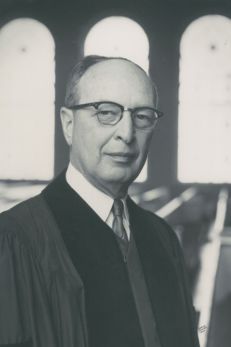 Historical black and white portrait of Rabbi Jacob Rothschild, an influential Jewish American rabbi and civil rights advocate. He is captured wearing his rabbinical robes, with a dignified and serene expression. The background features arched windows of a synagogue, symbolizing his deep connection to his faith and his community. This image underscores his role as a moral and spiritual leader during pivotal movements in American history.