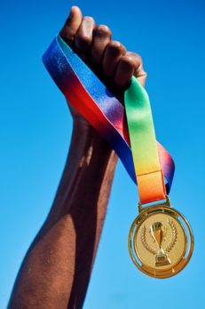 A hand is holding up a gold medal with a multicolored ribbon against a clear blue sky. The medal features a trophy symbol and laurel wreaths. The arm and hand are raised in a gesture of victory and celebration.