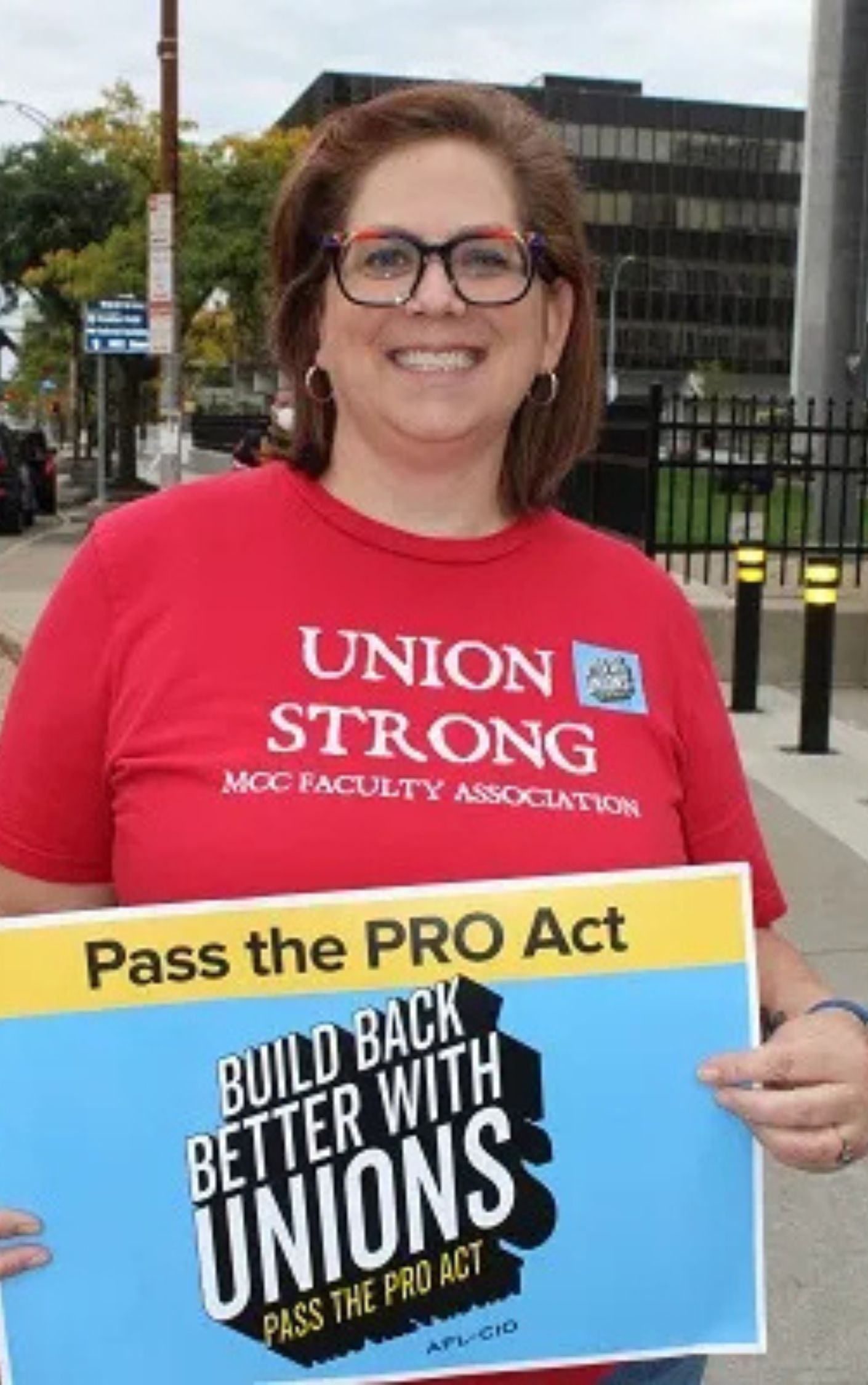 A smiling woman wearing a red "Union Strong – MCC Faculty Association" T-shirt holds a sign that reads "Pass the PRO Act – Build Back Better with Unions." She is standing outdoors on a city sidewalk.