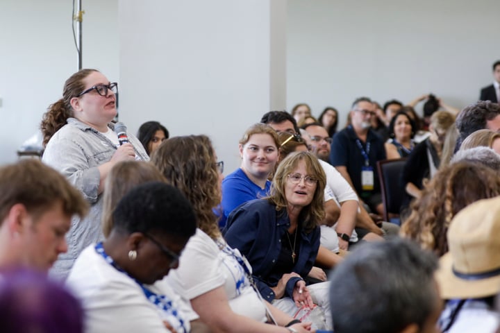 "Sarah Lerner speaks into a microphone during a packed session at the AFT TEACH 2025 conference. She stands among seated attendees, engaging with the panel while others listen attentively